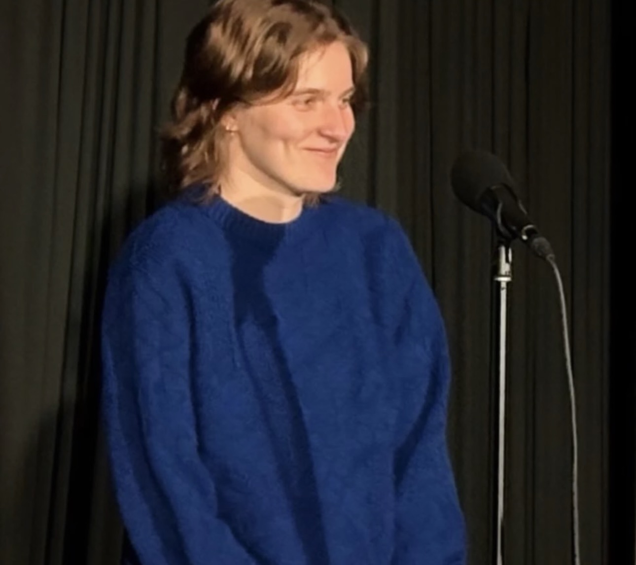 
                    A young woman (me) standing at a microphone in front of a black 
                    curtain. She is smiling and wearing a royal blue sweater.
                    