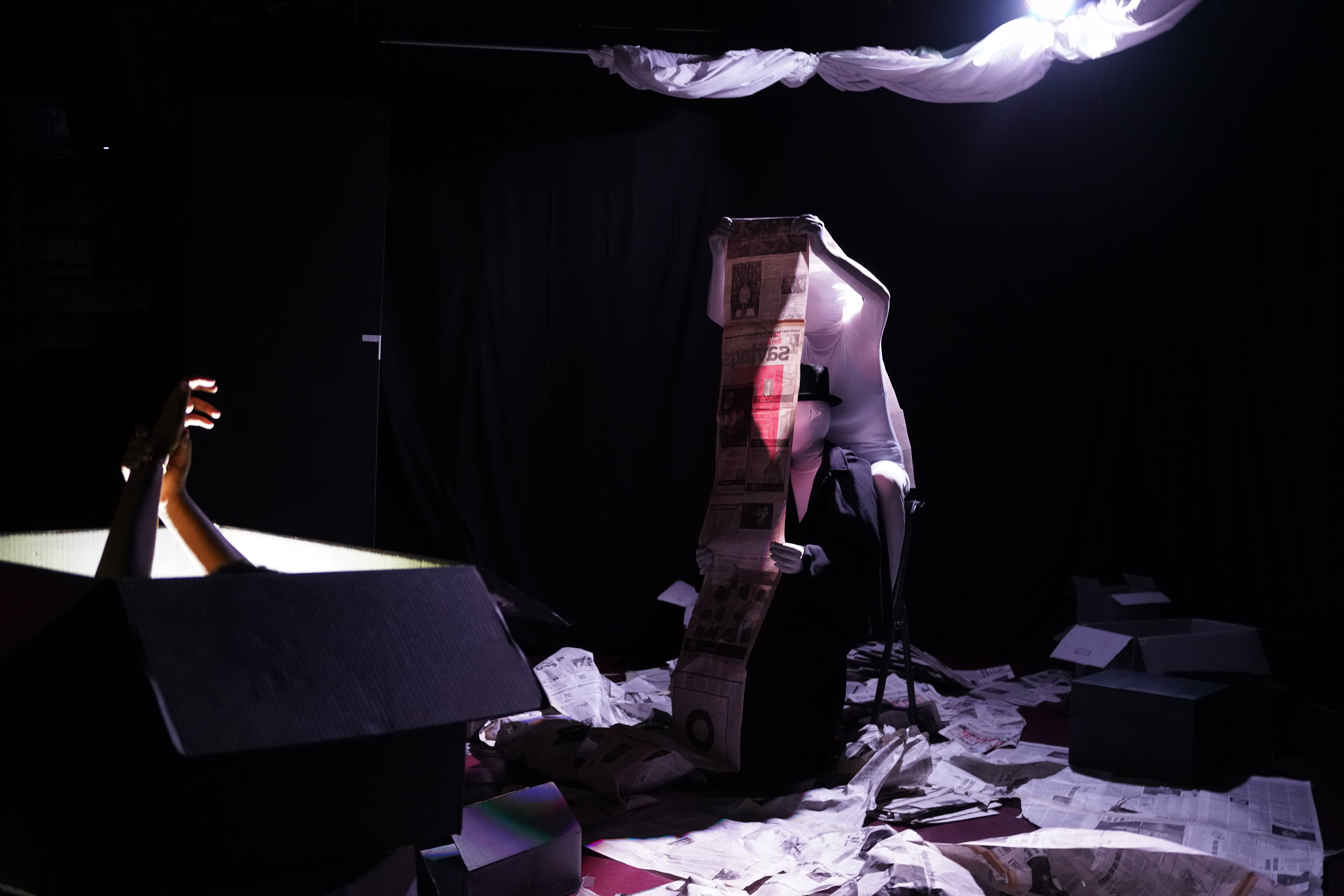 
          Anna in a white morphsuit, perched atop a chair. Sitting in the chair is another
          person in a white morphsuit and detective outfit. Together, they are reading a very
          long newspaper. There are newspapers covering the floor. In the foreground, two
          hairs emerge from a cardboard box. The box is lit from within.
          