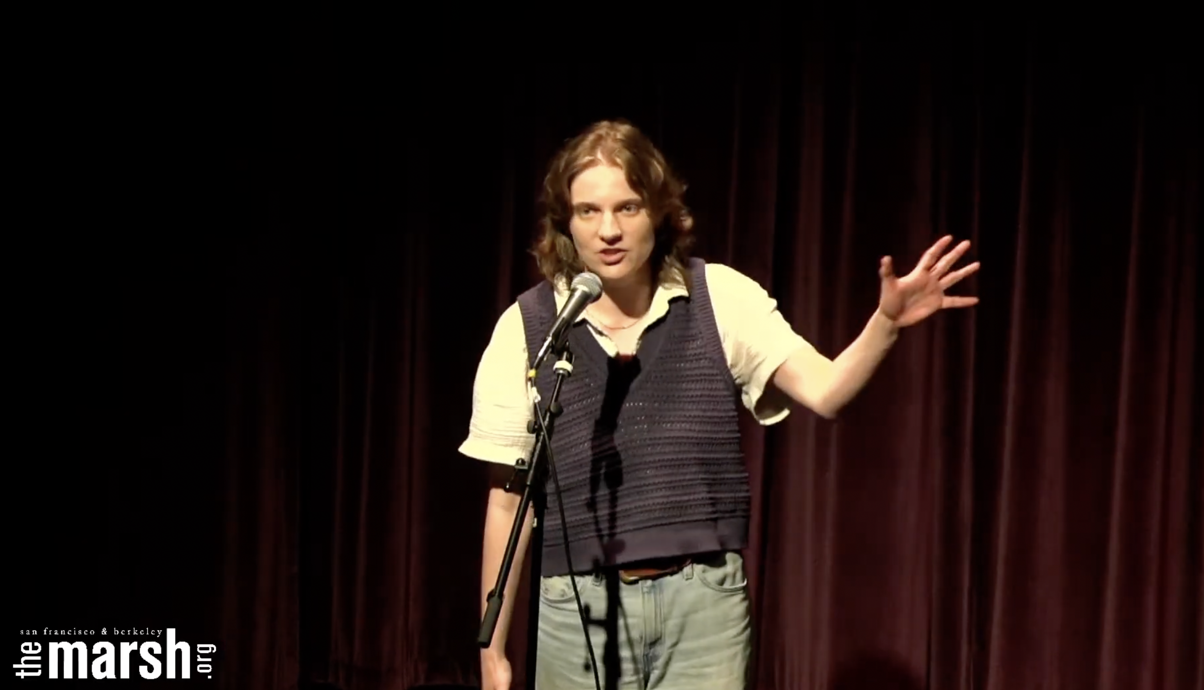 
                    A young woman (me) standing at a microphone in front of a dark red curtain. She is speaking and has her hand raised in an emphasizing gesture.
                    